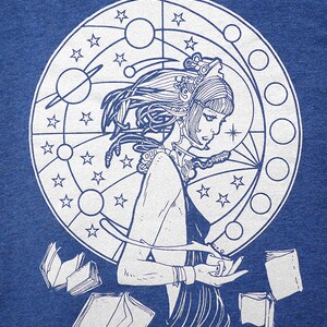 May include: A white graphic design on a blue t-shirt features a woman with long hair standing inside a circle with stars and planets. The woman is holding books in her hands.