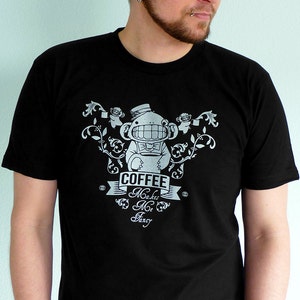Pu&ograve; includere: Una maglietta nera con un design grafico bianco di una scimmia dei cartoni animati che indossa un cappello a cilindro e tiene una tazza di caff&egrave;. Il testo "Coffee Makes Me Fancy" &egrave; stampato sulla maglietta.