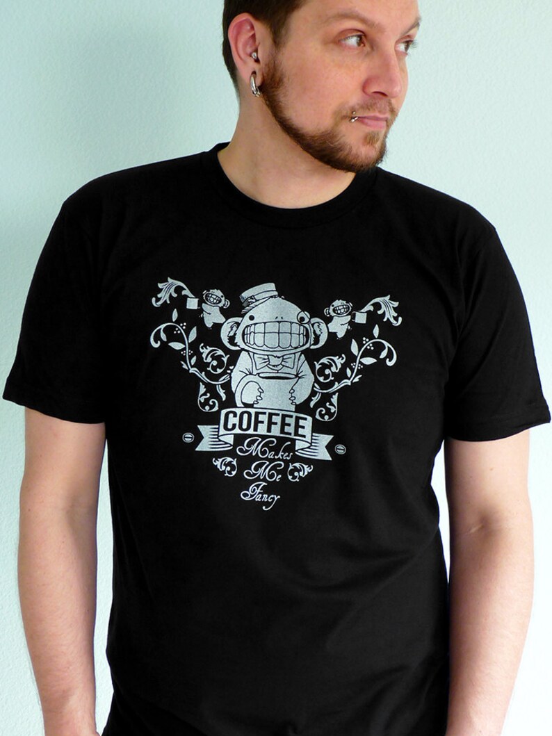 Pu&ograve; includere: Una maglietta nera con un design grafico bianco di una scimmia dei cartoni animati che indossa un cappello a cilindro e tiene una tazza di caff&egrave;. Il testo "Coffee Makes Me Fancy" &egrave; stampato sulla maglietta.