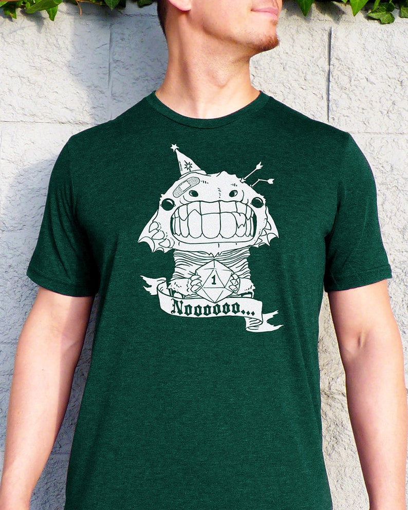 May include: A dark green t-shirt with a white graphic of a monster holding a dice with the number one on it. The monster is wearing a party hat and has a bandage on its head. The text "Nooooooooo..." is written on a banner below the monster.