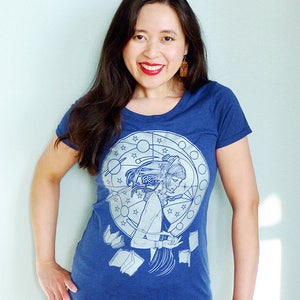 May include: A woman wearing a blue t-shirt with a white graphic print of a woman reading a book surrounded by stars and a circle. The graphic also includes the text "Library"