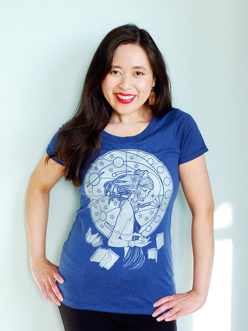 May include: A woman wearing a blue t-shirt with a white graphic print of a woman reading a book surrounded by stars and a circle. The graphic also includes the text "Library"