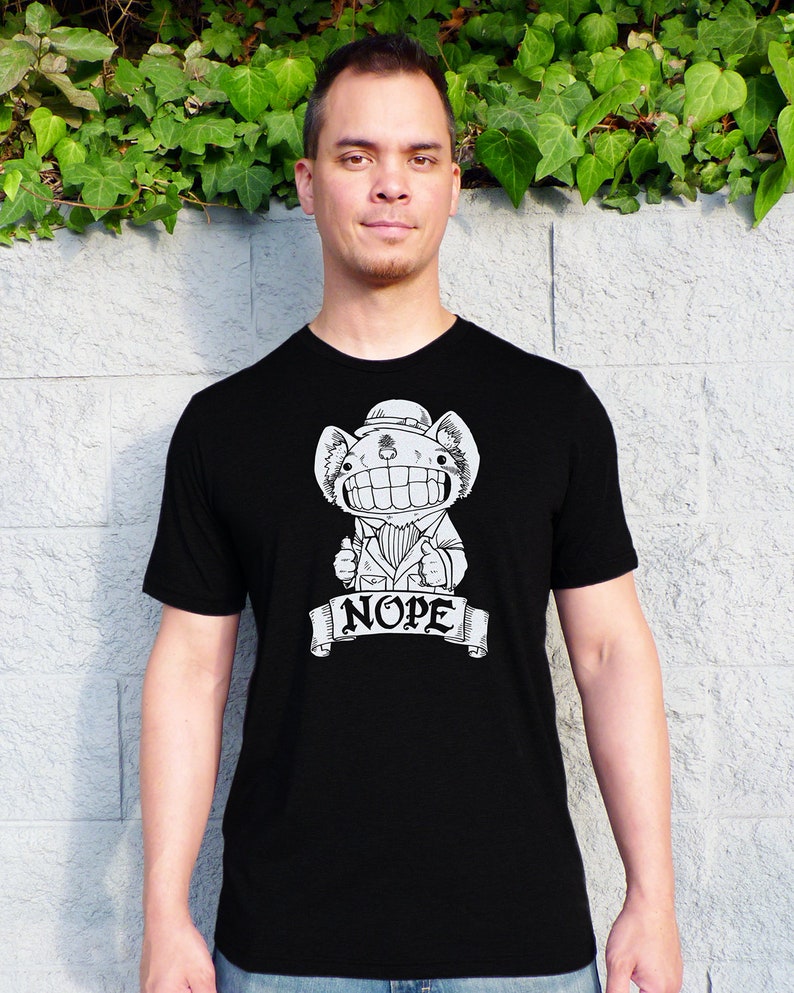 May include: A black t-shirt featuring a white graphic of a cartoon animal wearing a hat and a bow tie. The animal is giving a thumbs up and the text "NOPE" is written on a banner below it.