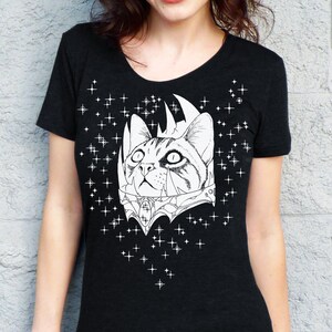 May include: Black t-shirt featuring a detailed white illustration of a cat with a crown and ornate collar. The cat is surrounded by a constellation of white stars. The shirt has a scoop neck and short sleeves.