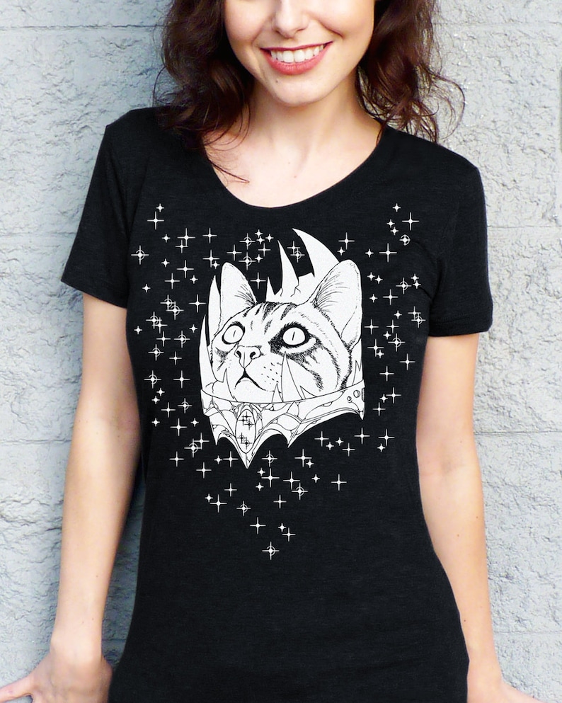 May include: Black t-shirt featuring a detailed white illustration of a cat with a crown and ornate collar. The cat is surrounded by a constellation of white stars. The shirt has a scoop neck and short sleeves.