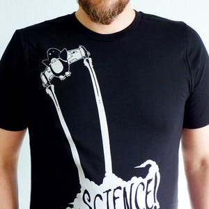 K&ouml;nnte beinhalten: Schwarzes T-Shirt mit einem wei&szlig;en Aufdruck eines Pinguins in einer Rakete, darunter das Wort "SCIENCE!". Das Design ist eine lustige, wissenschaftlich angehauchte Illustration. Der Pinguin wird in einer Rakete mit Abgasfahnen dargestellt.