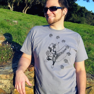 May include: A gray t-shirt with a black and white illustration of a squirrel holding an acorn. The squirrel is surrounded by acorns. The t-shirt has a crew neck and short sleeves.