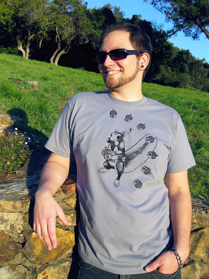 May include: A gray t-shirt with a black and white illustration of a squirrel holding an acorn. The squirrel is surrounded by acorns. The t-shirt has a crew neck and short sleeves.
