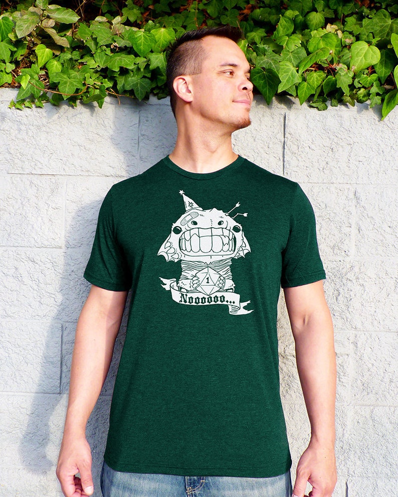 May include: A green t-shirt with a white graphic of a cartoon monster holding a die with the number one on it. The monster is wearing a party hat and has a banner that says 'Nooooooooo...'.