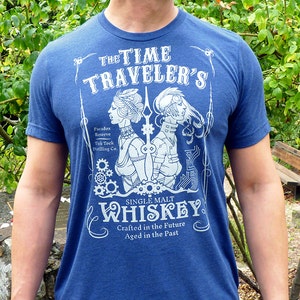 May include: A blue t-shirt with a white graphic design of a vintage whiskey label. The label features a stylized illustration of two figures with gears and the text "THE TIME TRAVELER'S SINGLE MALT WHISKEY Crafted in the Future Aged in the Past".