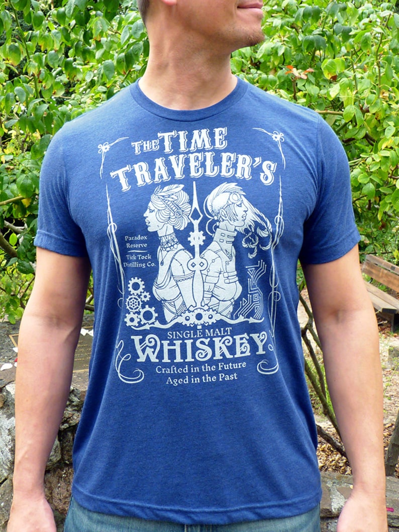 May include: A blue t-shirt with a white graphic design of a vintage whiskey label. The label features a stylized illustration of two figures with gears and the text "THE TIME TRAVELER'S SINGLE MALT WHISKEY Crafted in the Future Aged in the Past".