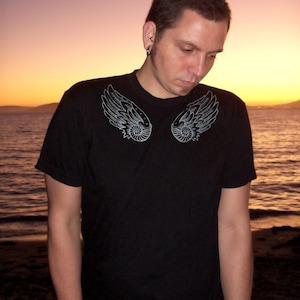 May include: A black t-shirt with a white design of two stylized wings. The wings have a detailed pattern of circles and lines.