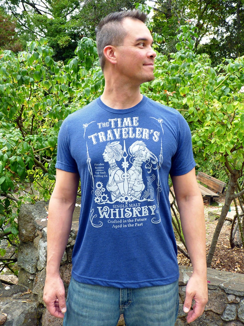 May include: A blue t-shirt with a white graphic design that reads 'The Time Traveler's' and 'Single Malt Whiskey Crafted in the Future Aged in the Past' with a vintage style illustration of two women with gears and a clock.