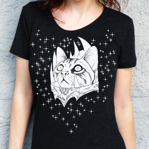 May include: Black t-shirt featuring a detailed black and white illustration of a cat wearing a crown and ornate collar. White star graphics are scattered around the cat design. The shirt has a scoop neck.