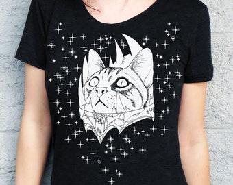 May include: Black t-shirt featuring a detailed black and white illustration of a cat wearing a crown and ornate collar. White star graphics are scattered around the cat design. The shirt has a scoop neck.