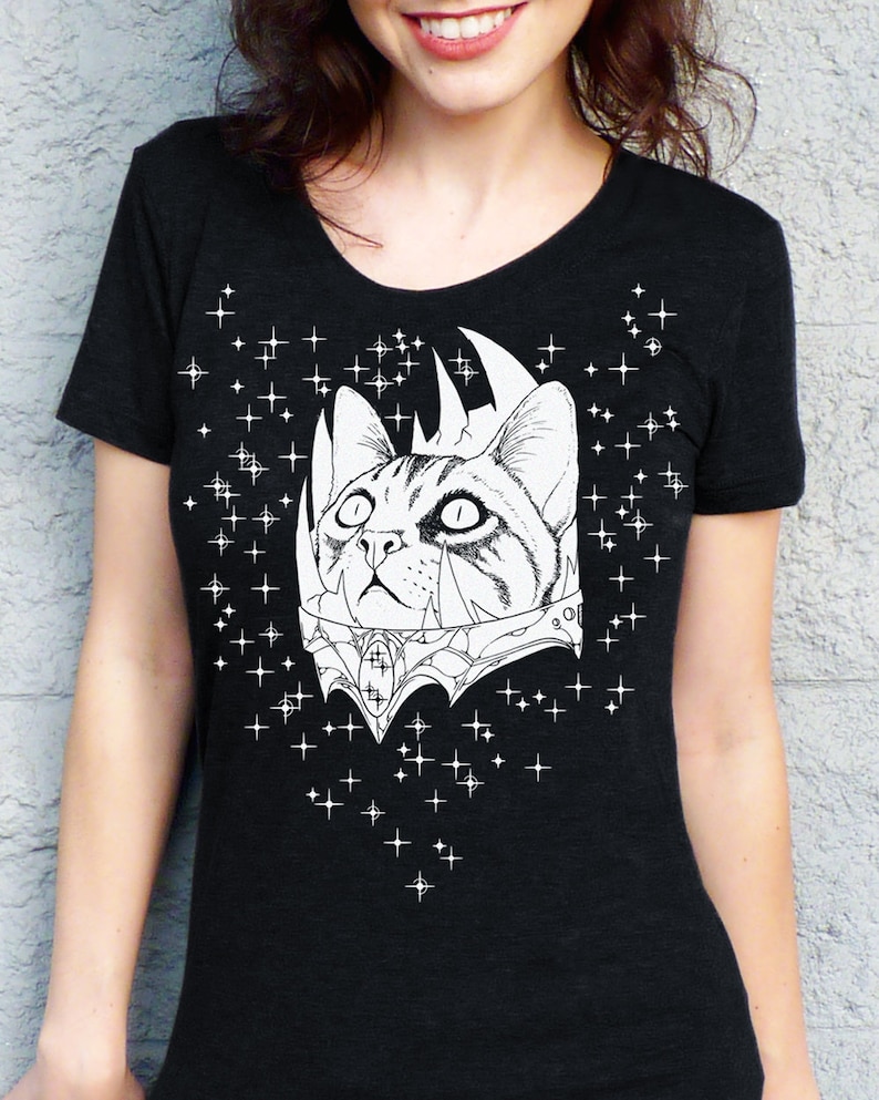 May include: Black t-shirt featuring a detailed black and white illustration of a cat wearing a crown and ornate collar. White star graphics are scattered around the cat design. The shirt has a scoop neck.