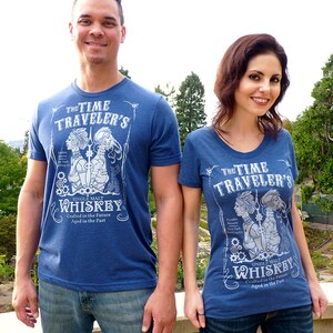 May include: Two people wearing blue t-shirts with a white graphic design featuring a vintage style illustration of a couple with the text "The Time Traveler's Single Malt Whiskey Crafted in the Future Aged in the Past".