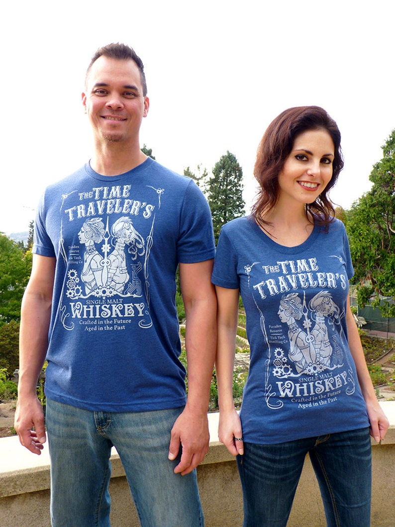 May include: Two people wearing blue t-shirts with a white graphic design featuring a vintage style illustration of a couple with the text "The Time Traveler's Single Malt Whiskey Crafted in the Future Aged in the Past".