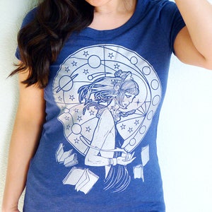 May include: A navy blue t-shirt with a white graphic print of a woman with long hair, surrounded by stars, planets, and books. The graphic is in a circular design.