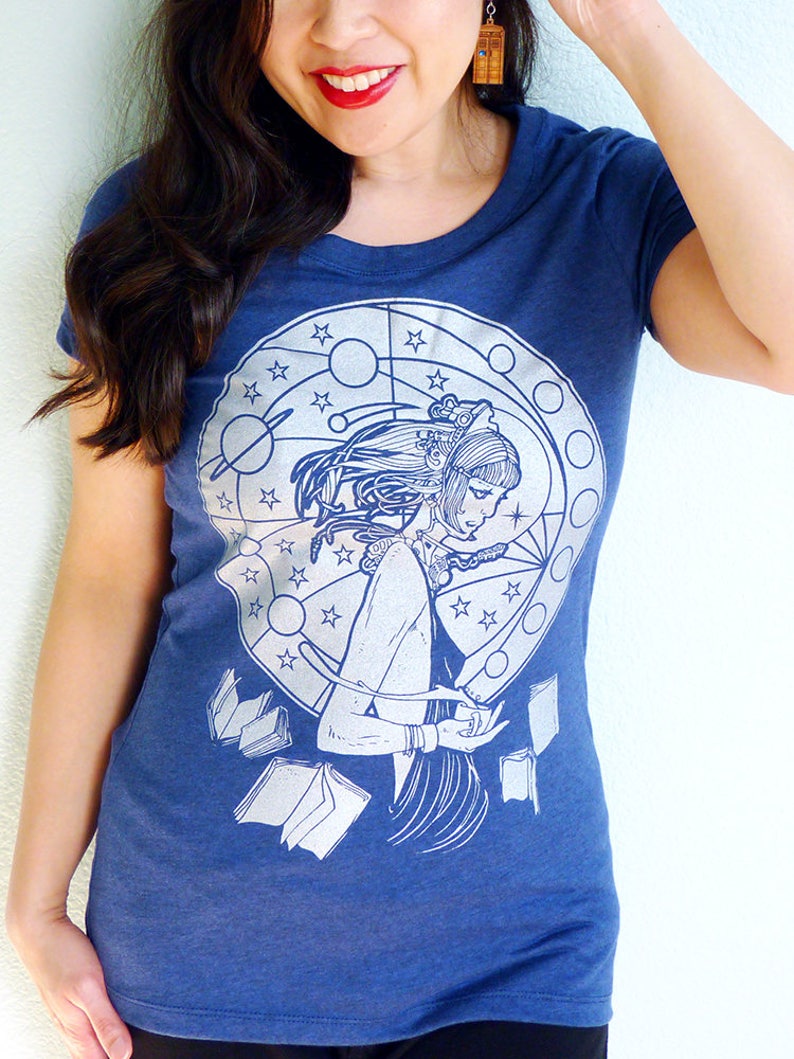 May include: A navy blue t-shirt with a white graphic print of a woman with long hair, surrounded by stars, planets, and books. The graphic is in a circular design.