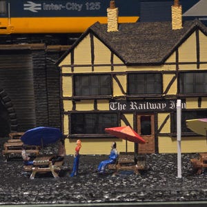 Può includere: Un modello in miniatura di un treno giallo e blu che passa sopra un ponte con un piccolo edificio giallo a due piani con finiture nere e il testo "The Railway Inn" sopra l'ingresso. L'edificio ha un tetto nero e diverse finestre. Ci sono persone in miniatura e ombrelloni davanti all'edificio.