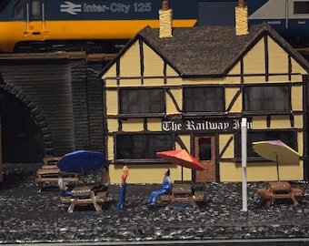 May include: A miniature model of a yellow and blue train passing over a bridge with a small, two-story, yellow building with black trim and the text "The Railway Inn" above the entrance. The building has a black roof and several windows. There are miniature people and umbrellas in front of the building.