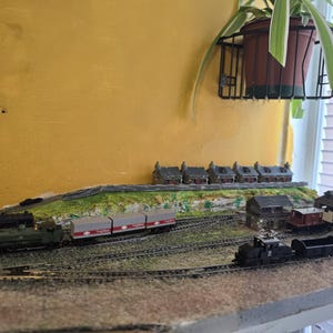 May include: A model train set featuring a black locomotive pulling a line of grey boxcars. The train is on a track that runs through a miniature landscape with small houses and a stone building.