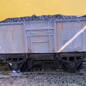 May include: A model train coal car, painted in shades of gray with a white diagonal stripe. The car is filled with black coal and sits on a model railroad track. The number "0972" is visible on the side.