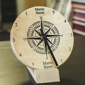 May include: A circular light-coloured wooden tide clock featuring a black compass rose graphic in the centre. The face displays numbers 1 through 5 on both sides, with the text Marée Haute at the top and Marée Basse at the bottom, and black hands.