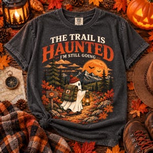 May include: A charcoal gray short-sleeve t-shirt featuring a graphic of a ghost wearing a red beanie and carrying a green backpack while holding a lantern on a forest path. The text reads THE TRAIL IS HAUNTED I'M STILL GOING in orange and white.