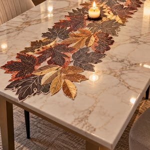 Pode incluir: Um caminho de mesa com um arranjo intrincado de folhas de bordo bordadas em tons de castanho, laranja e dourado. O caminho está colocado sobre uma mesa de mármore branco, com o texto TheSeedsGrowIn visível no canto superior esquerdo.