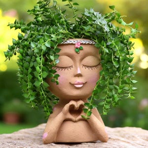 May include: A tan-colored resin planter shaped like a human head with closed eyes, pink cheeks, and a small pink flower detail on a silver headband. The figure has hands positioned in a heart shape at the chest, filled with trailing green foliage.