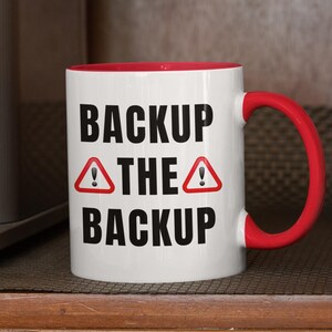 Peut inclure: Mug en céramique blanche avec intérieur et anse rouges. Le mug affiche le texte "BACKUP THE BACKUP" en noir, avec des symboles d'avertissement rouges. Le mug est placé à côté d'une machine à café.