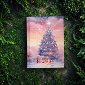 May include: A hardcover journal featuring a winter scene with a decorated Christmas tree. The illustration has a soft, watercolor style with a pink and purple sky, snow, and a small house. The journal is surrounded by green foliage.