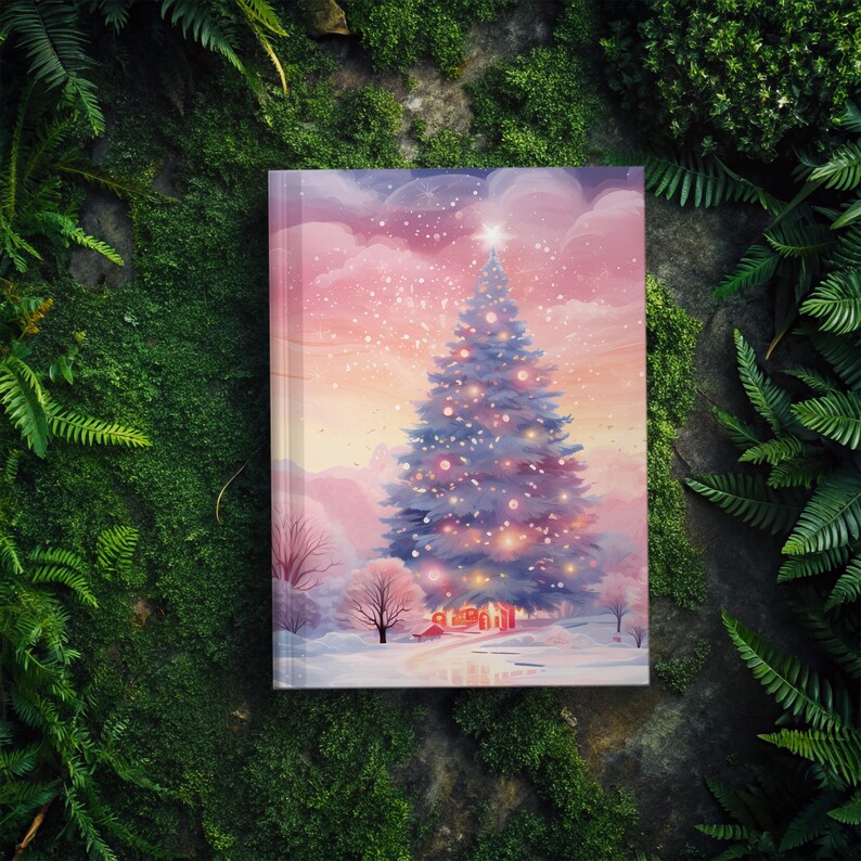 May include: A hardcover journal featuring a winter scene with a decorated Christmas tree. The illustration has a soft, watercolor style with a pink and purple sky, snow, and a small house. The journal is surrounded by green foliage.
