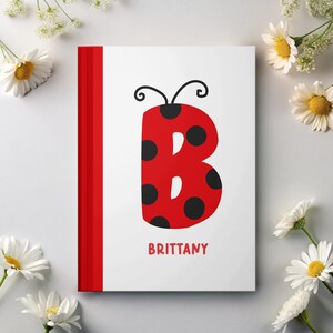 May include: A white notebook with a red spine and the letter "B" designed as a ladybug. The ladybug is red with black spots and black antennae. The name "BRITTANY" is printed in red below the letter.