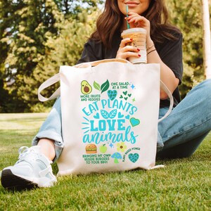 May include: White canvas tote bag with the text "Eat Plants Love Animals" in blue and green. The bag also features a green avocado, a green apple, a burger, a mushroom, and hearts.