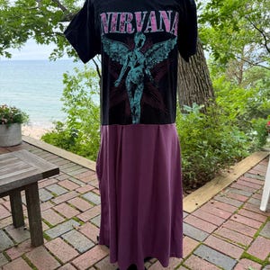 May include: A black t-shirt dress featuring a graphic print of the word NIRVANA in pink letters above a teal winged figure. The upper portion is a black short-sleeved shirt, while the lower portion is a solid dark purple skirt with pleats.