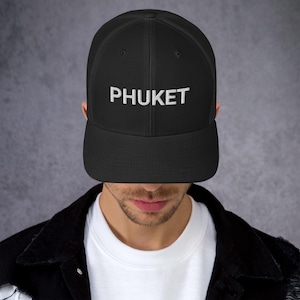 May include: A black trucker-style baseball cap featuring a structured front panel with the word PHUKET embroidered in white capital letters. The cap includes a solid black brim and a mesh back panel, providing a classic look with a flat visor.