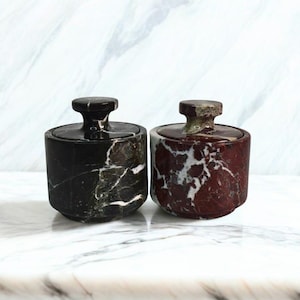 May include: Two marble jars with lids. One jar is black with white veining, and the other is a deep red with white veining. The jars have a cylindrical shape with a small, round knob on top of the lid.
