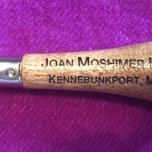 May include: A wooden crochet hook with a blue metal hook and the text "JOAN MOSHIMER HOOK KENNEBUNKPORT, MAINE" engraved on the handle.
