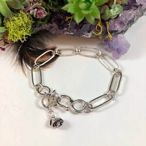 Op de afbeelding: Een zilveren kettingarmband met een roosbedel. De armband is gemaakt van ovale schakels en heeft een karabijn sluiting.