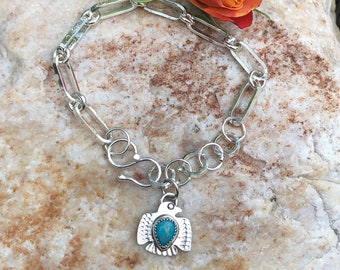 Puede incluir: Un brazalete de cadena de plata con un dije de águila de turquesa y plata. El brazalete está sobre un fondo de roca con una flor.