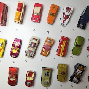 Puede incluir: Una colección de coches de juguete vintage, que incluyen un autobús de dos pisos rojo, un camión de bomberos amarillo, un coche deportivo verde y una camioneta azul. Los coches están dispuestos en filas sobre una superficie blanca.