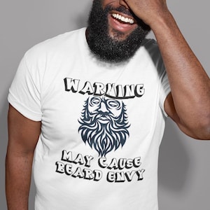 May include: White t-shirt with the text "WARNING MAY CAUSE BEARD ENVY" and a stylized beard graphic. The person wearing the shirt is smiling and covering their eyes with their hand.