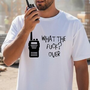 May include: White t-shirt with a black graphic of a walkie-talkie and the text "WHAT THE FUCK? OVER." The shirt is short-sleeved and worn by a person wearing a yellow hard hat and holding a walkie-talkie.