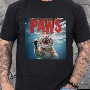 May include: Black t-shirt featuring a graphic of a cat underwater with the word "PAWS" in red above it. The cat is orange and white, with its mouth open and a paw raised. The shirt is a classic crew neck.