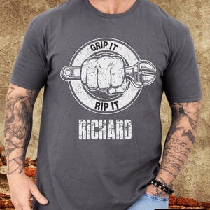 May include: Charcoal gray t-shirt with a white graphic of a fist gripping a wrench inside a circle, with the words "GRIP IT" and "RIP IT" above and below. The name "RICHARD" is printed below the graphic.