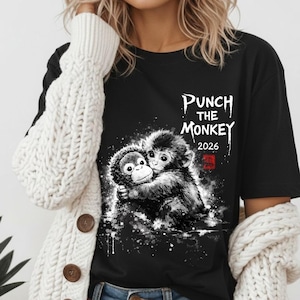 May include: Black t-shirt featuring a grayscale illustration of two hugging monkeys. The text "PUNCH THE MONKEY 2026" is printed above the image, with a red square symbol below. The design has a distressed, artistic style.