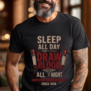 May include: A black short-sleeved t-shirt featuring the text SLEEP ALL DAY DRAW BLOOD ALL NIGHT CERTIFIED PHLEBOTOMIST SINCE 2026. The graphic includes two blood collection tubes, a syringe, and a caduceus symbol in red, white, and tan.